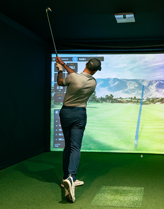 Golfer swinging a club inside a home simulator with a virtual course displayed on the screen.