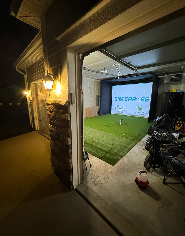 Wide shot from outside an open garage at night showing a completed home golf simulator inside.