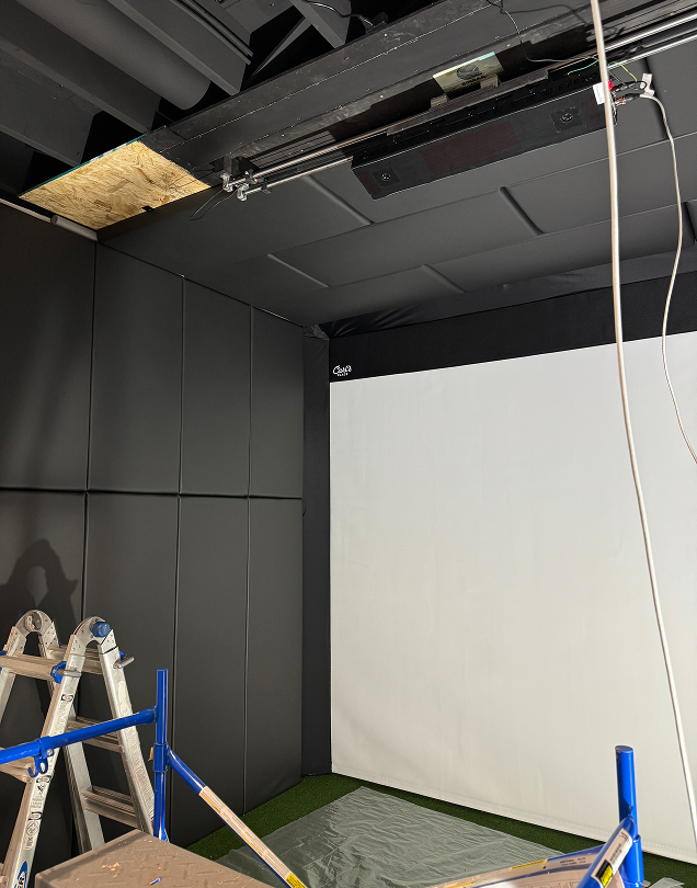 Construction view of a home golf simulator setup showing ceiling tracks, wall padding, and a blank white screen.